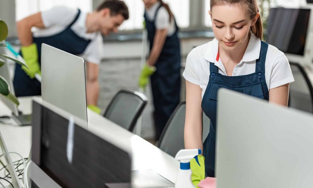 selective-focus-of-pretty-cleaner-wiping-desk-while-colleagues-cleaning-floor-in-office.jpg